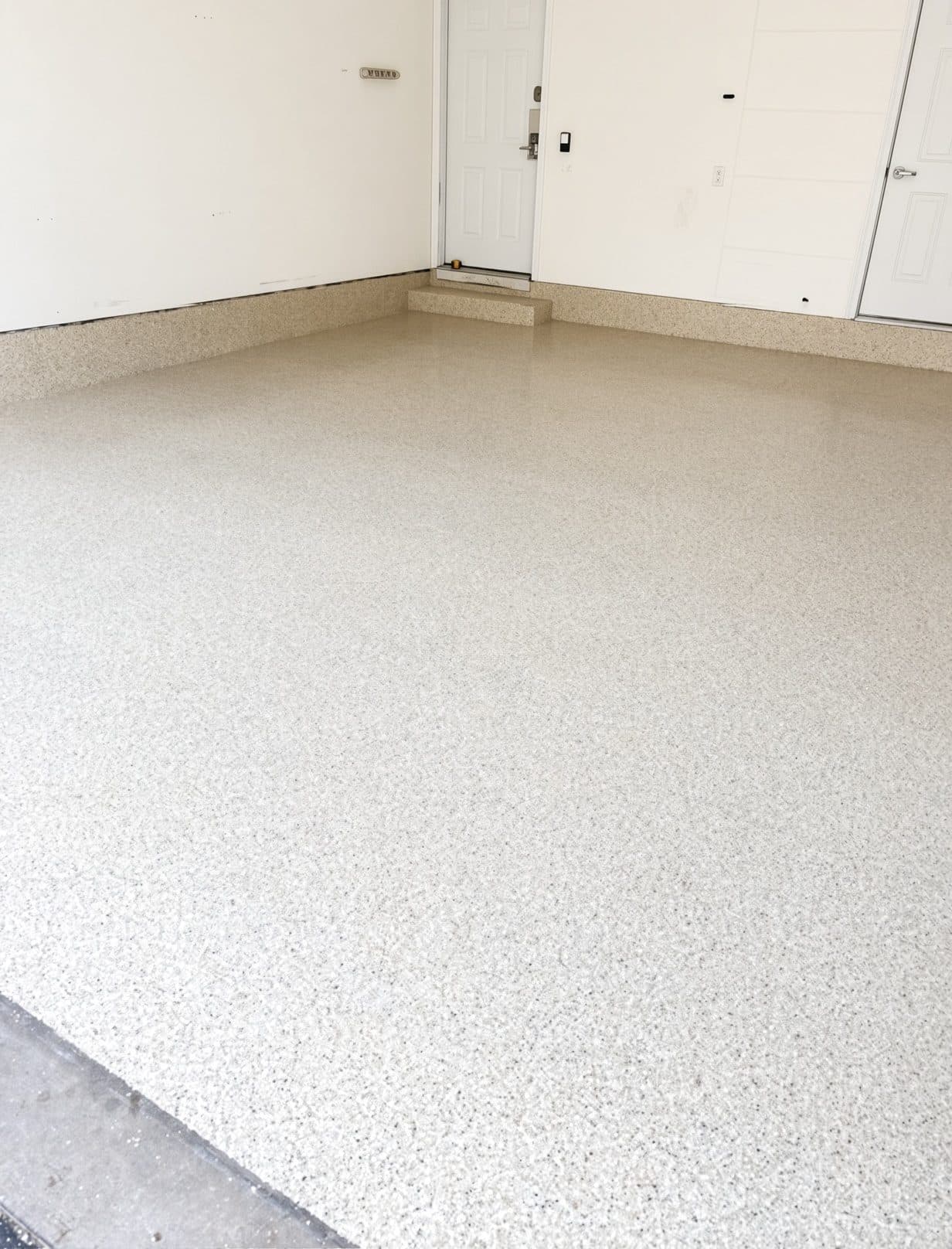 The same garage floor after a full-flake coating — an even neutral flake finish, seamless across the slab, carried up the stem walls and over the step, with a clean edge at the door.