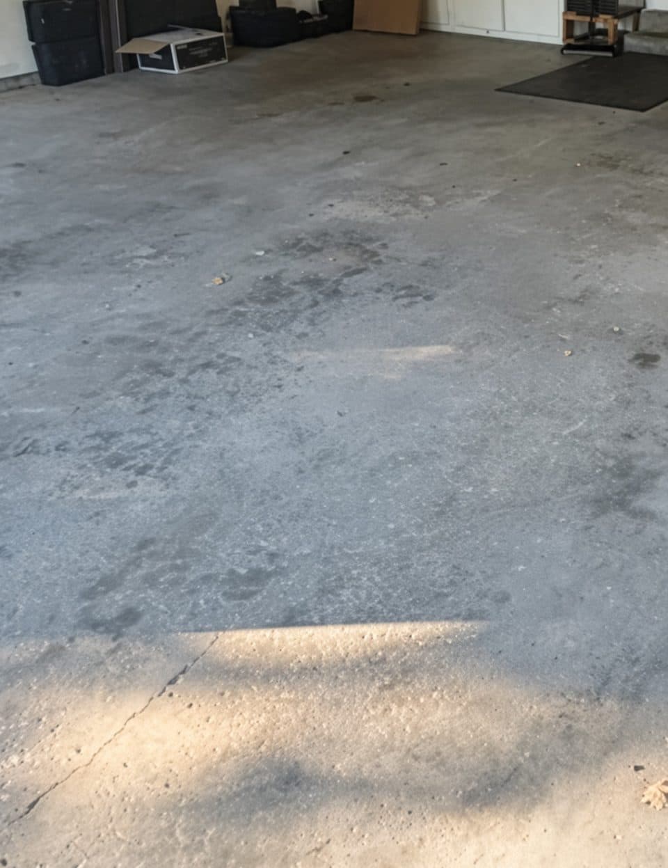 A two-car garage floor before coating — bare grey concrete, patchy and discolored, with a long crack running toward the door opening.